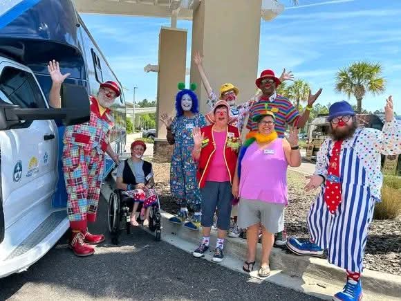 Group of people dressed as clowns and a person in a wheelchair outdoors, waving and smiling, with a vehicle and palm trees in background.
