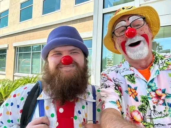 Two people dressed as clowns with red noses, smiling outdoors in front of a building, one with a hat and glasses.