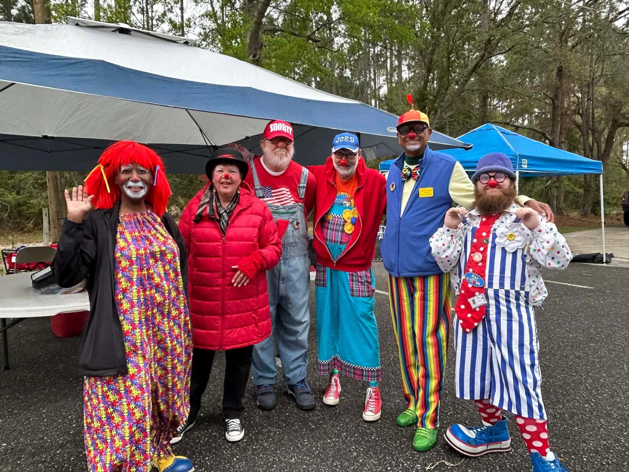 Group of six people dressed as clowns and circus performers standing outdoors under umbrellas, smiling and posing for the photo.