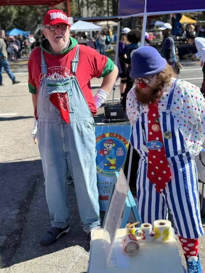 Two clowns in colorful costumes at an outdoor event, with tents and people in the background.