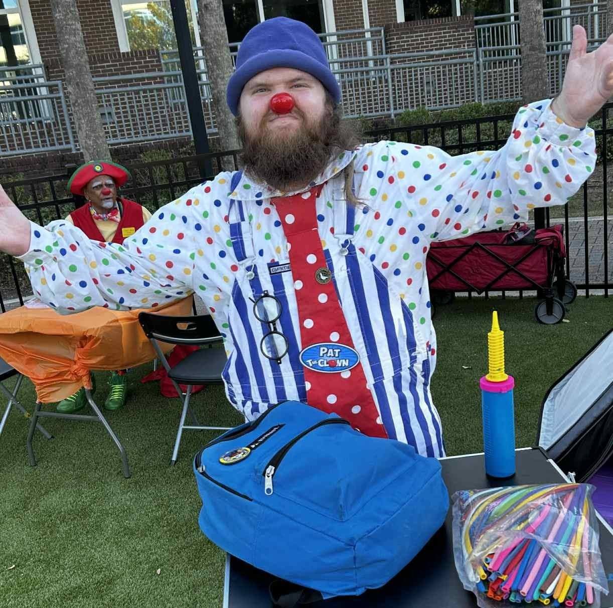 Person dressed as a clown with a red nose, blue hat, and colorful striped shirt, outdoors with a fence in background.