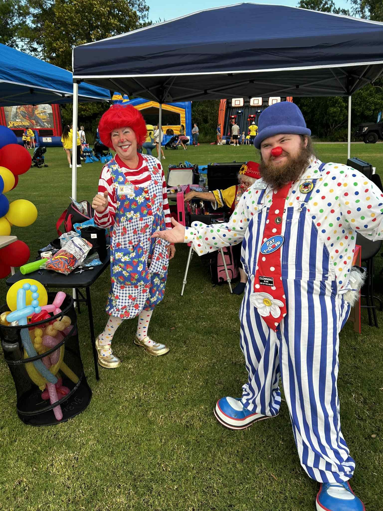 Two clowns in colorful costumes shake hands at an outdoor event under tents, with balloons and a playground in background.