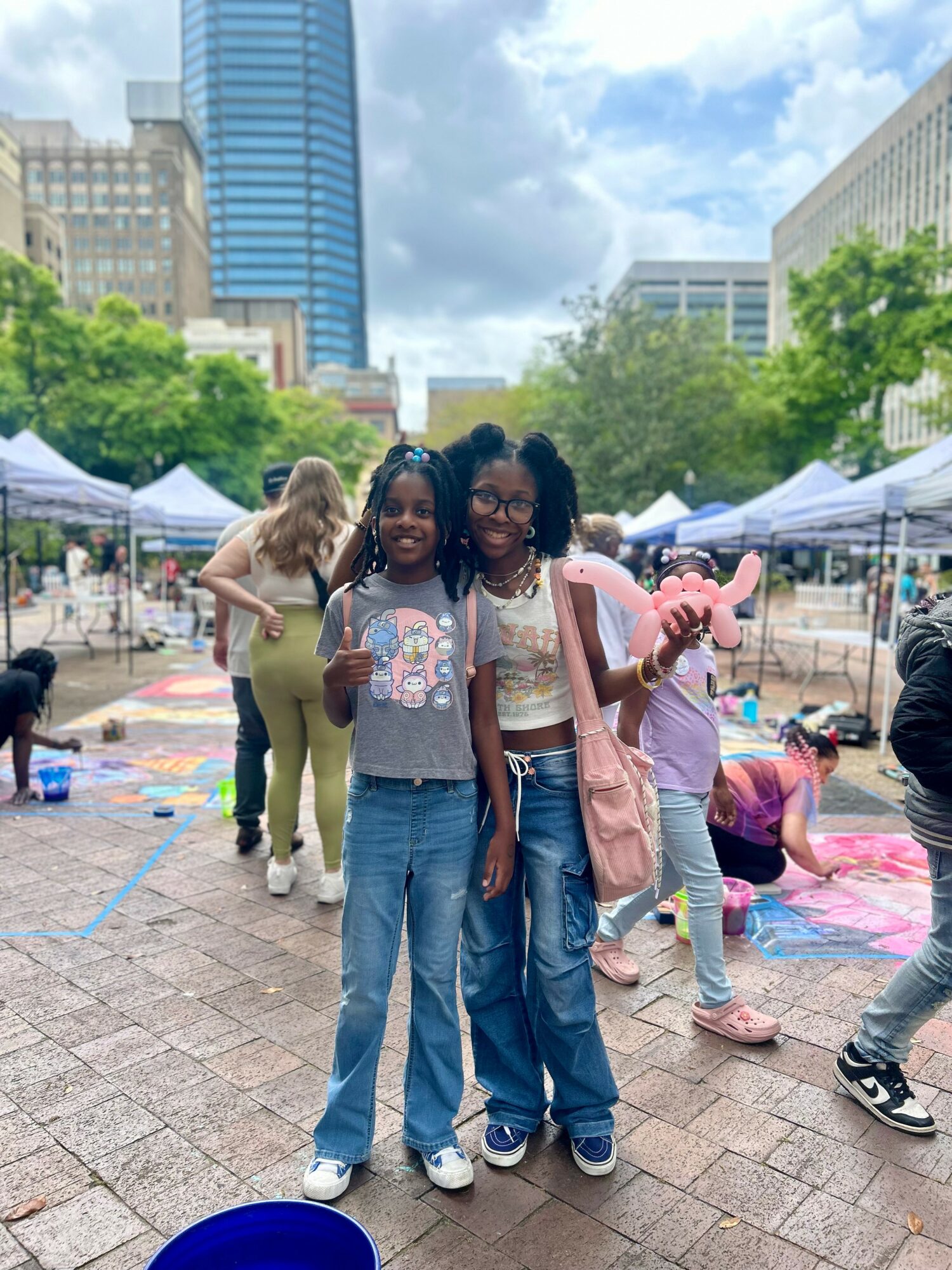 Two girls stand together smiling at an outdoor event with tents and people in the background.