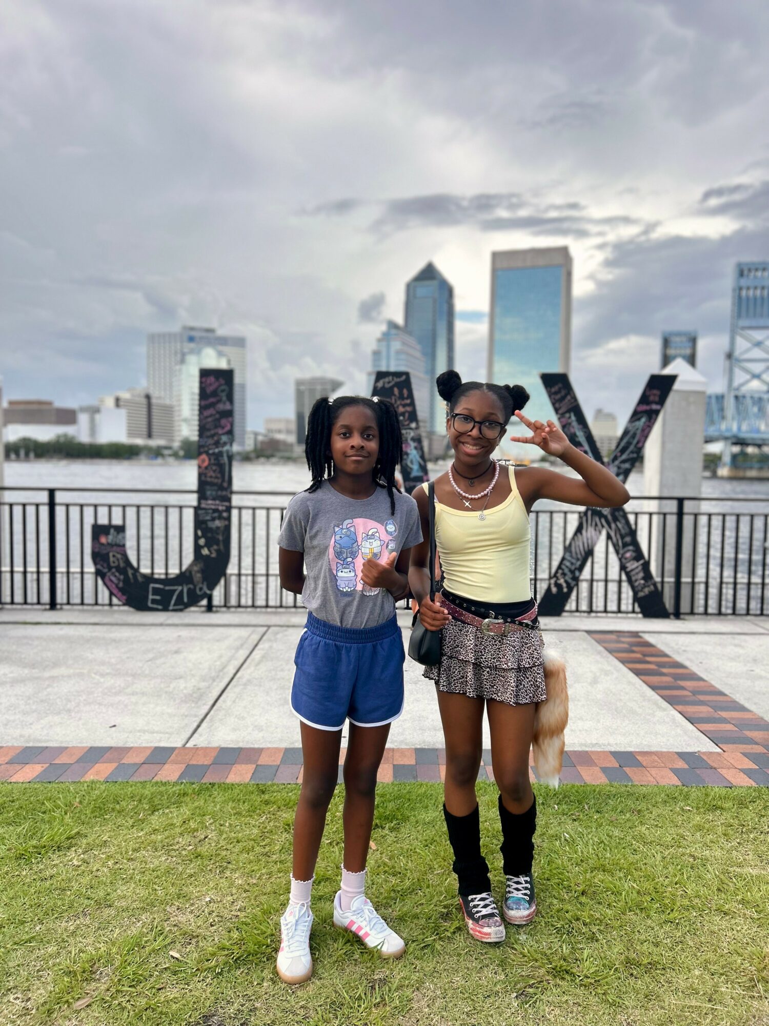 Two young women stand on a grassy area with city skyline and large letters behind them, one making a peace sign.