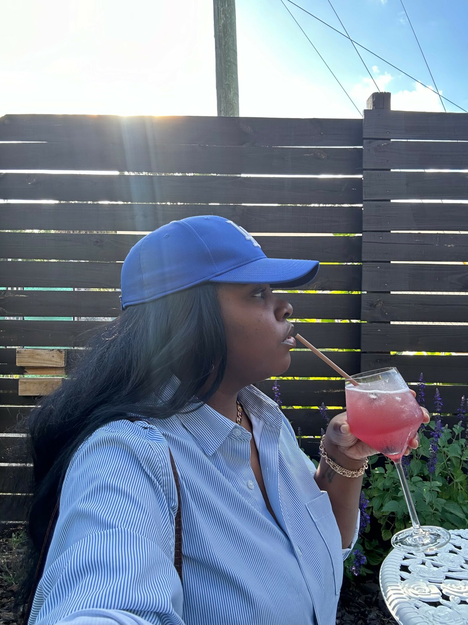 Woman in blue cap and striped shirt drinking pink beverage through a straw outdoors, wooden fence in background.
