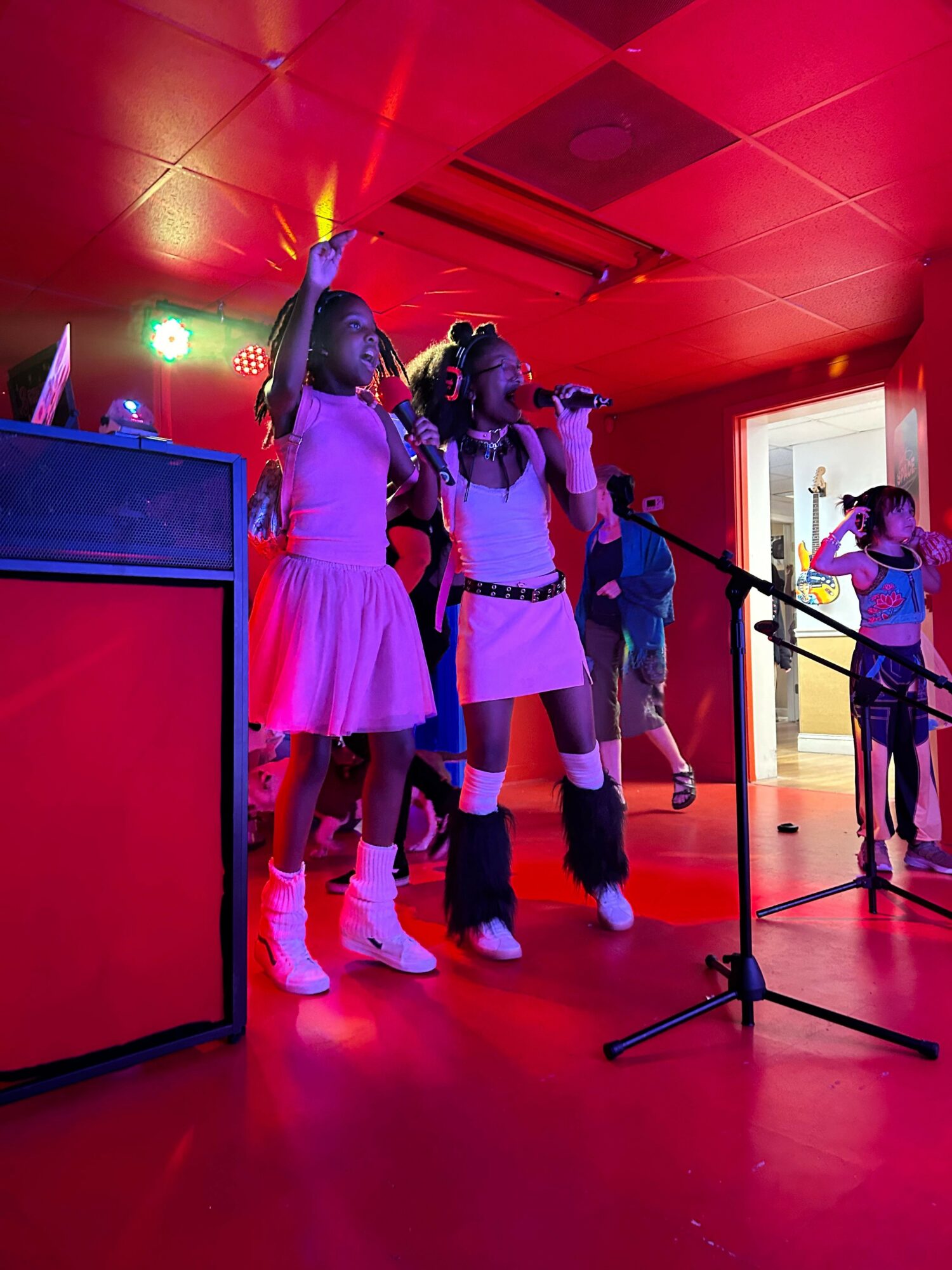 Group of children in a red hallway, some holding microphones, with a window and a person in the background.