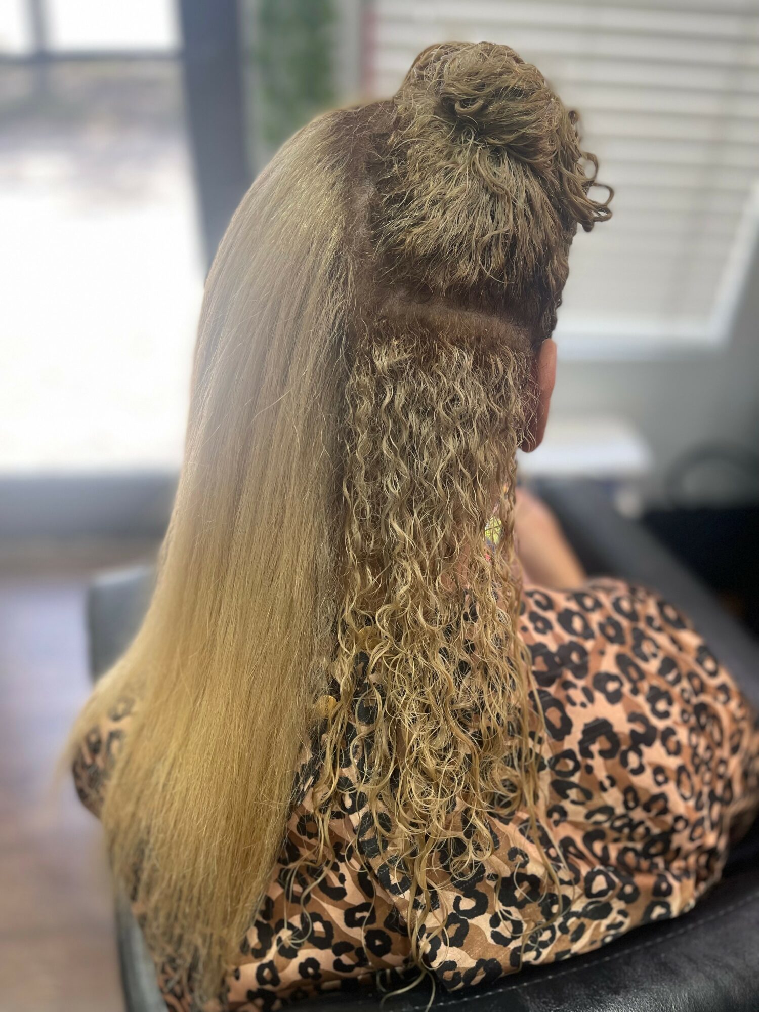 Person with long, curly blonde hair sitting indoors, back view, wearing a leopard print top.