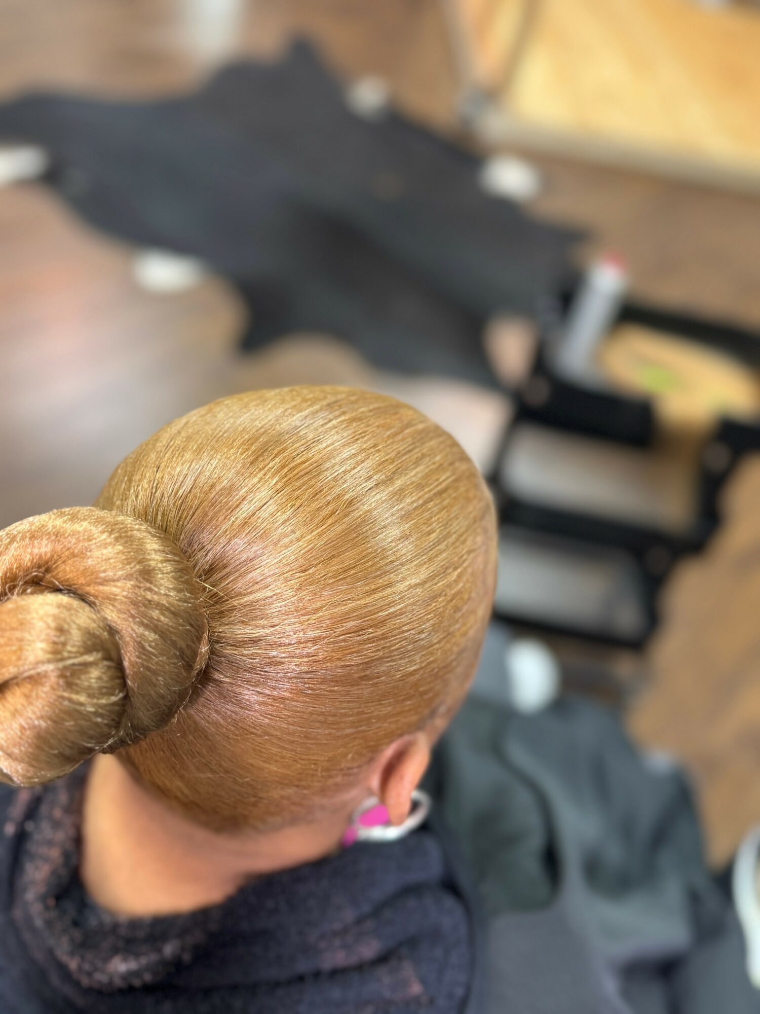 Top view of a person with blonde hair in a bun, sitting in a salon chair, with a blurred background of salon equipment.