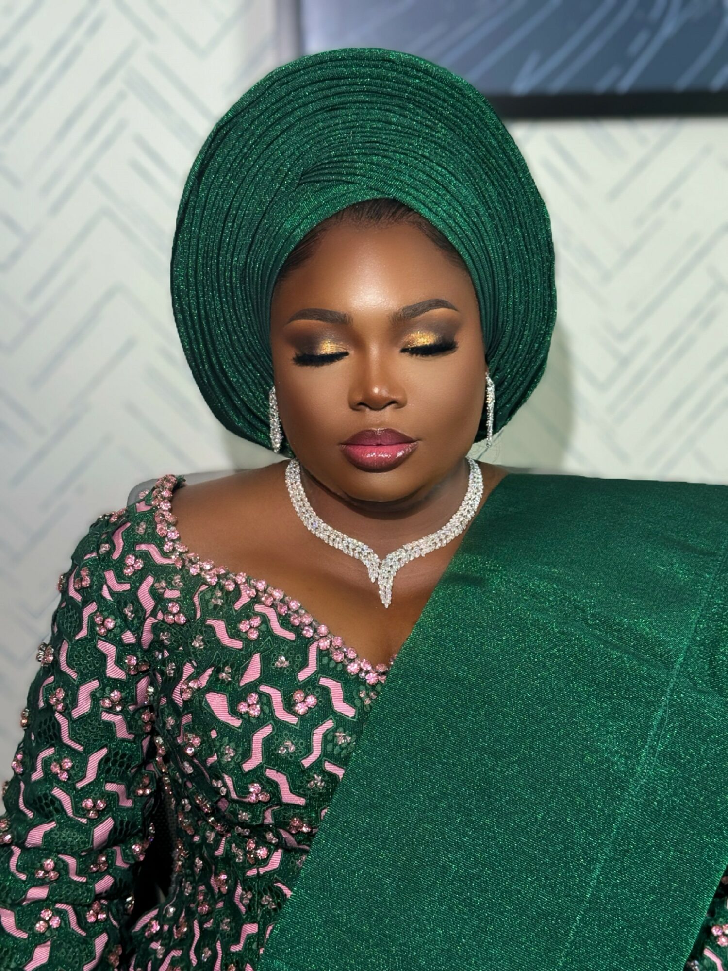 Woman with green headwrap and traditional attire, wearing jewelry, with eyes closed, in front of patterned background.