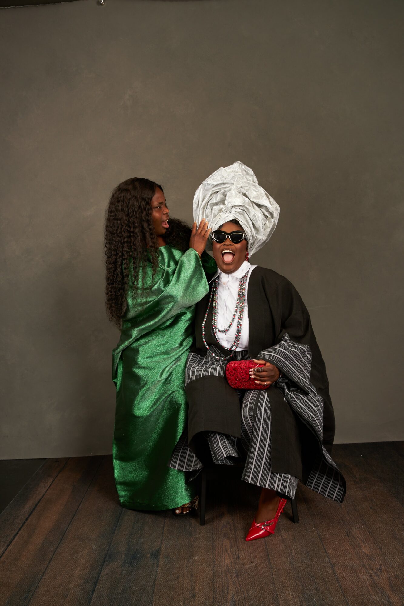Two women in traditional and modern attire pose together against a plain background, one wearing a green dress and the other in black with a headwrap.