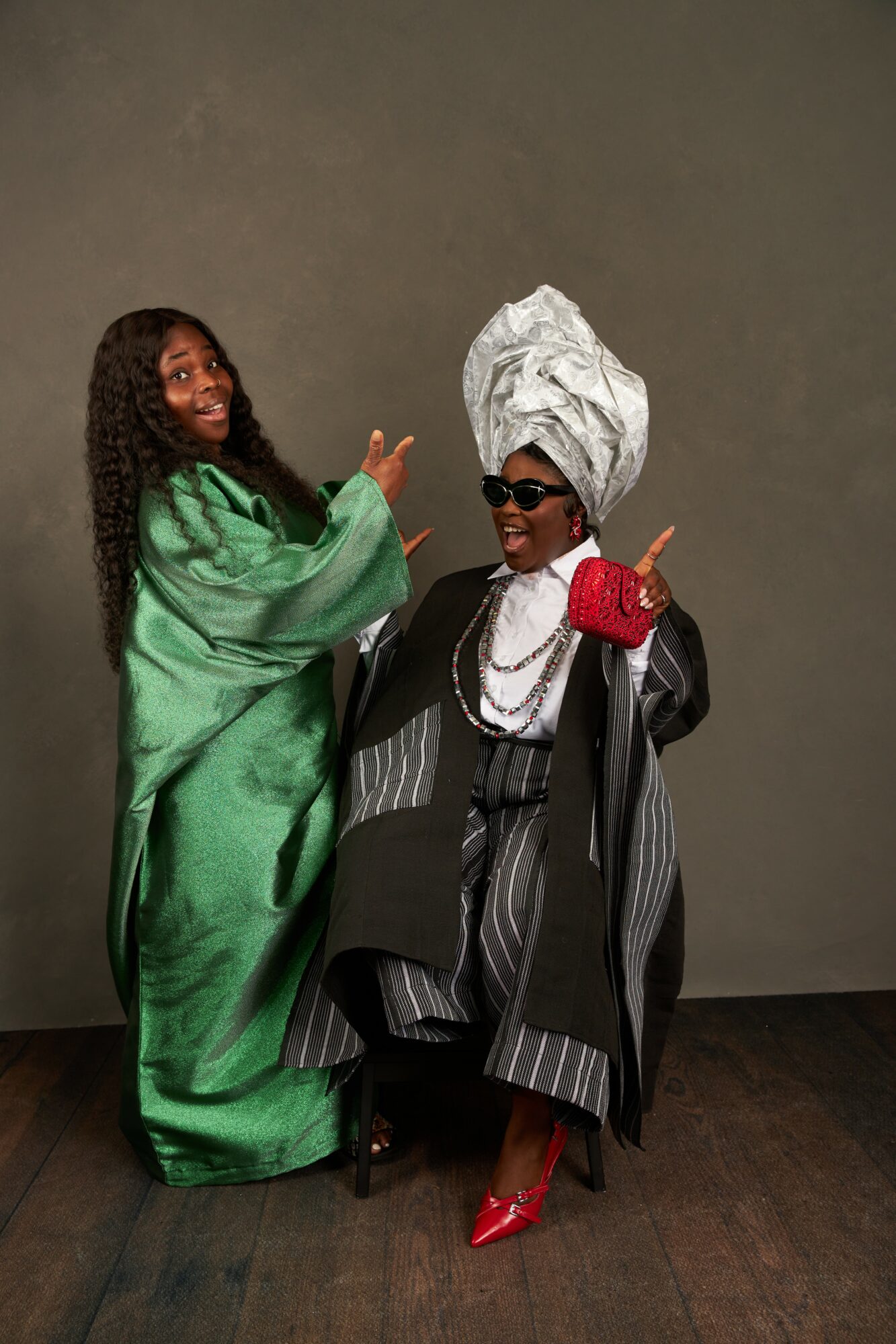 Two women pose together, one in a green dress and the other in a striped outfit with a large headwrap, sunglasses, and red shoes.