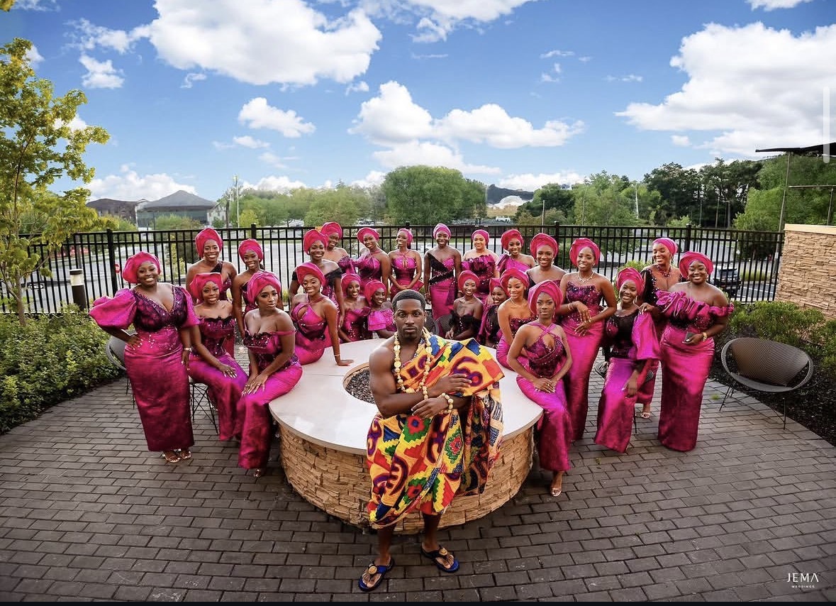 Group of women in pink traditional attire with one woman in colorful dress, outdoors on brick patio, with trees and sky.