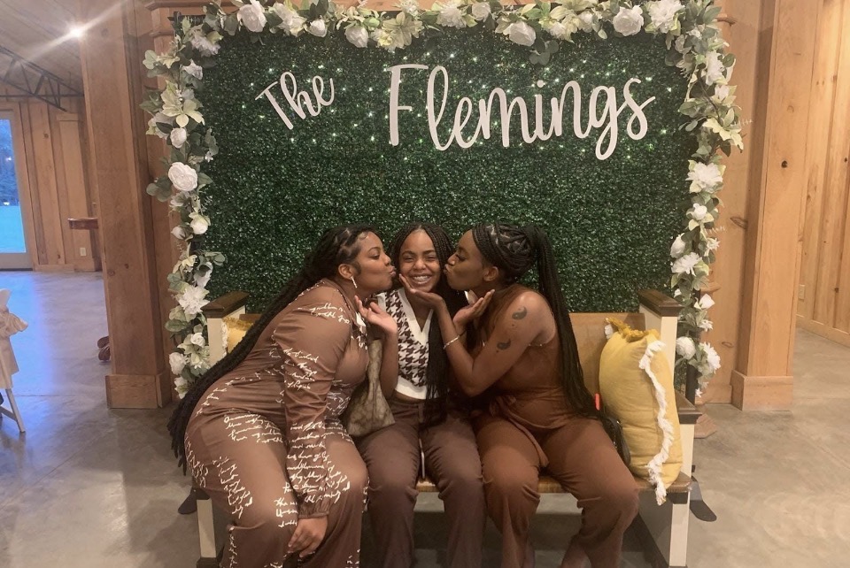 Three women sitting on a bench in front of a floral backdrop with the words 'The Flemings' written on it, smiling and posing together.