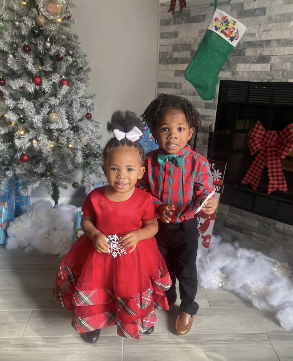 Two children in festive clothing stand in front of a decorated Christmas tree and fireplace with a stocking hanging above.