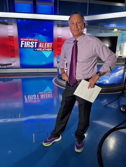 Male news anchor in studio holding papers, standing in front of weather graphics, wearing a light purple shirt and dark pants.