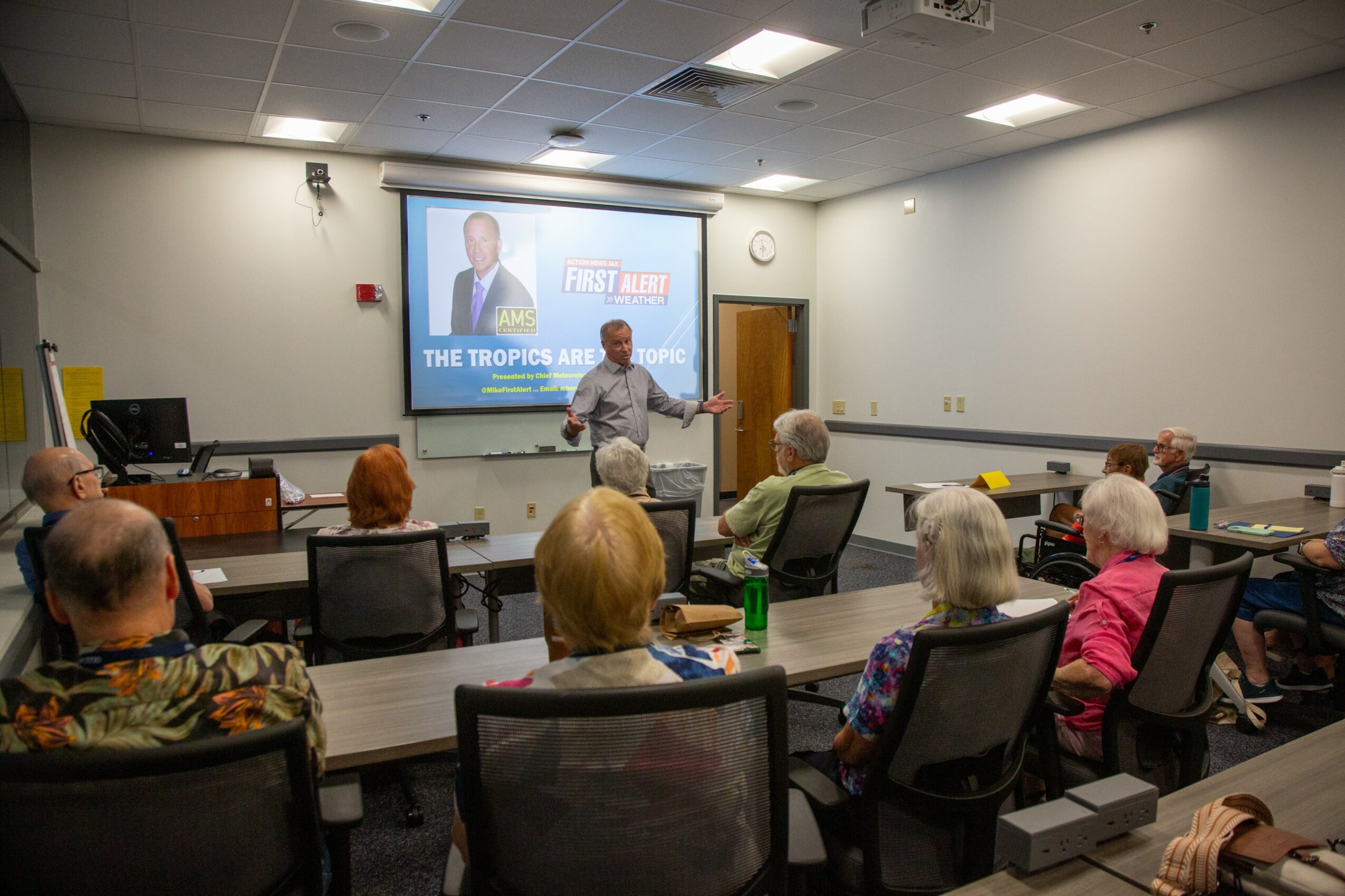 A man stands at the front of a classroom giving a presentation to seated adults, with a projected slide behind him.
