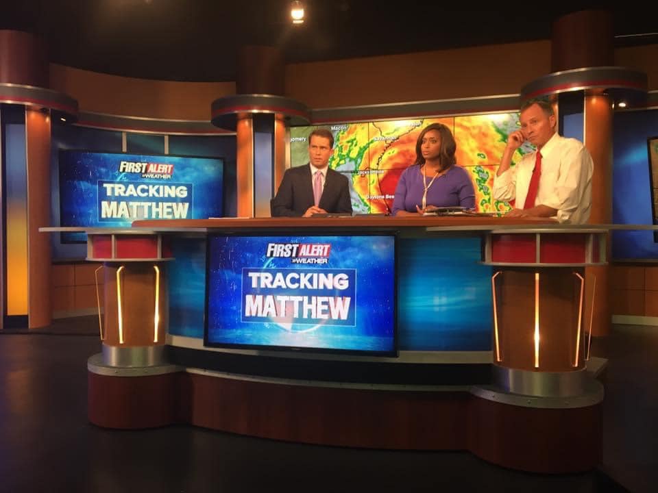 Three news anchors sit behind a desk in a television studio with a colorful background and a screen displaying weather graphics.
