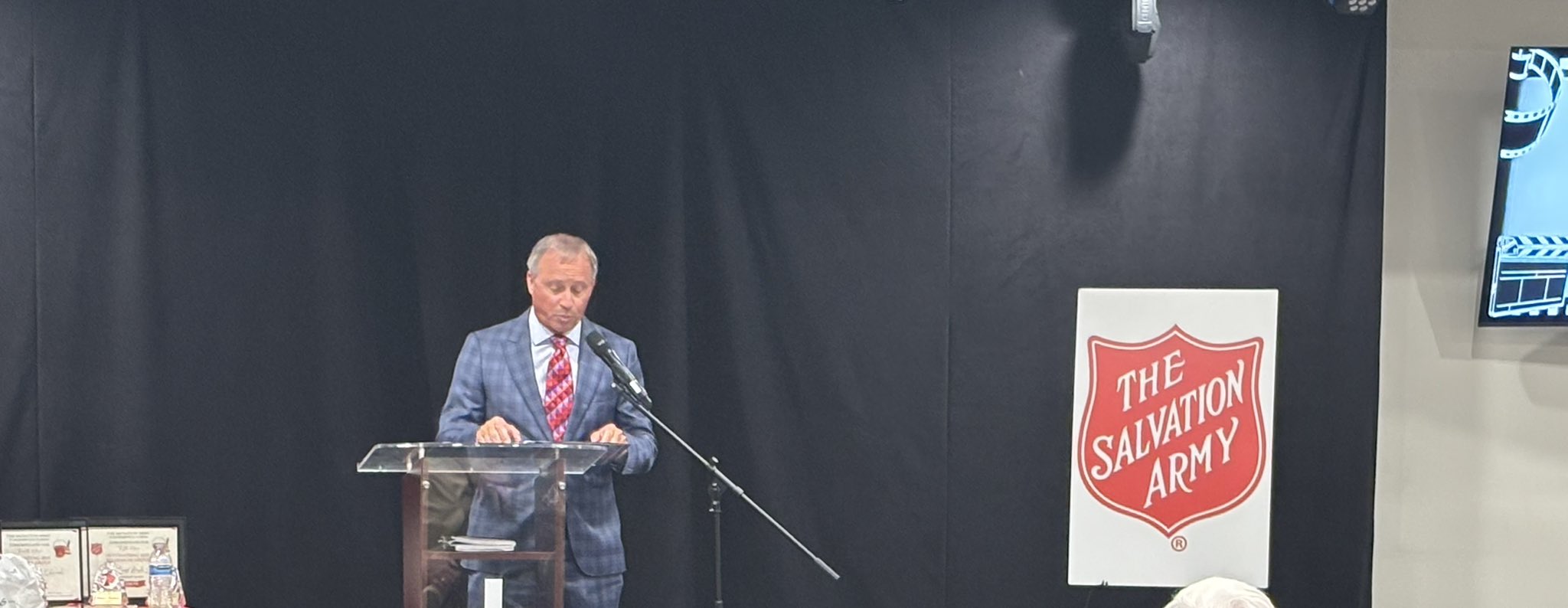 A man in a suit stands behind a clear podium on a stage with a black background and a Salvation Army sign.