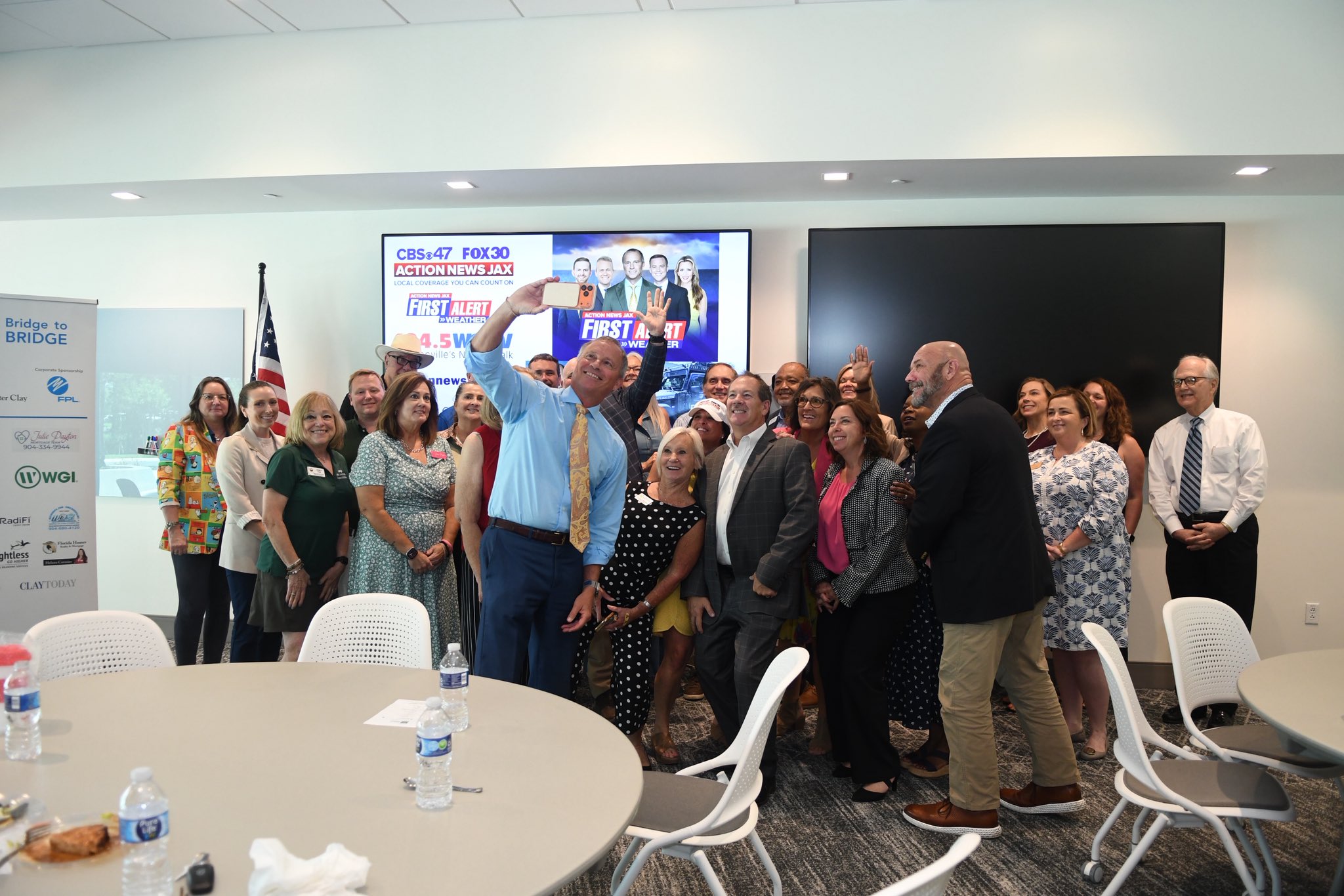 Group of people gathered in a room, some taking a selfie, with tables and chairs in the foreground.