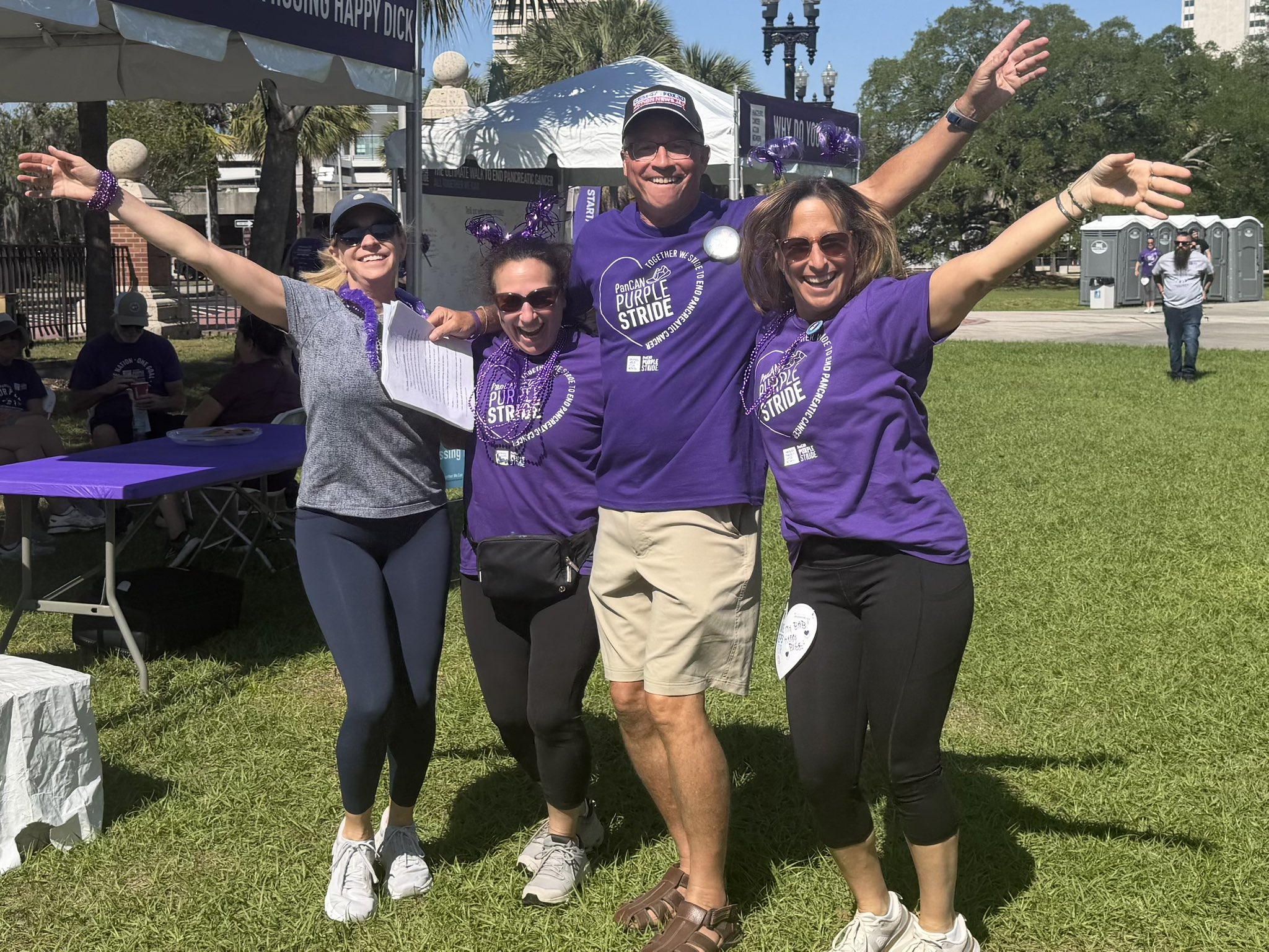 Four people in purple shirts standing outdoors with arms raised, smiling, on a grassy area during daytime.