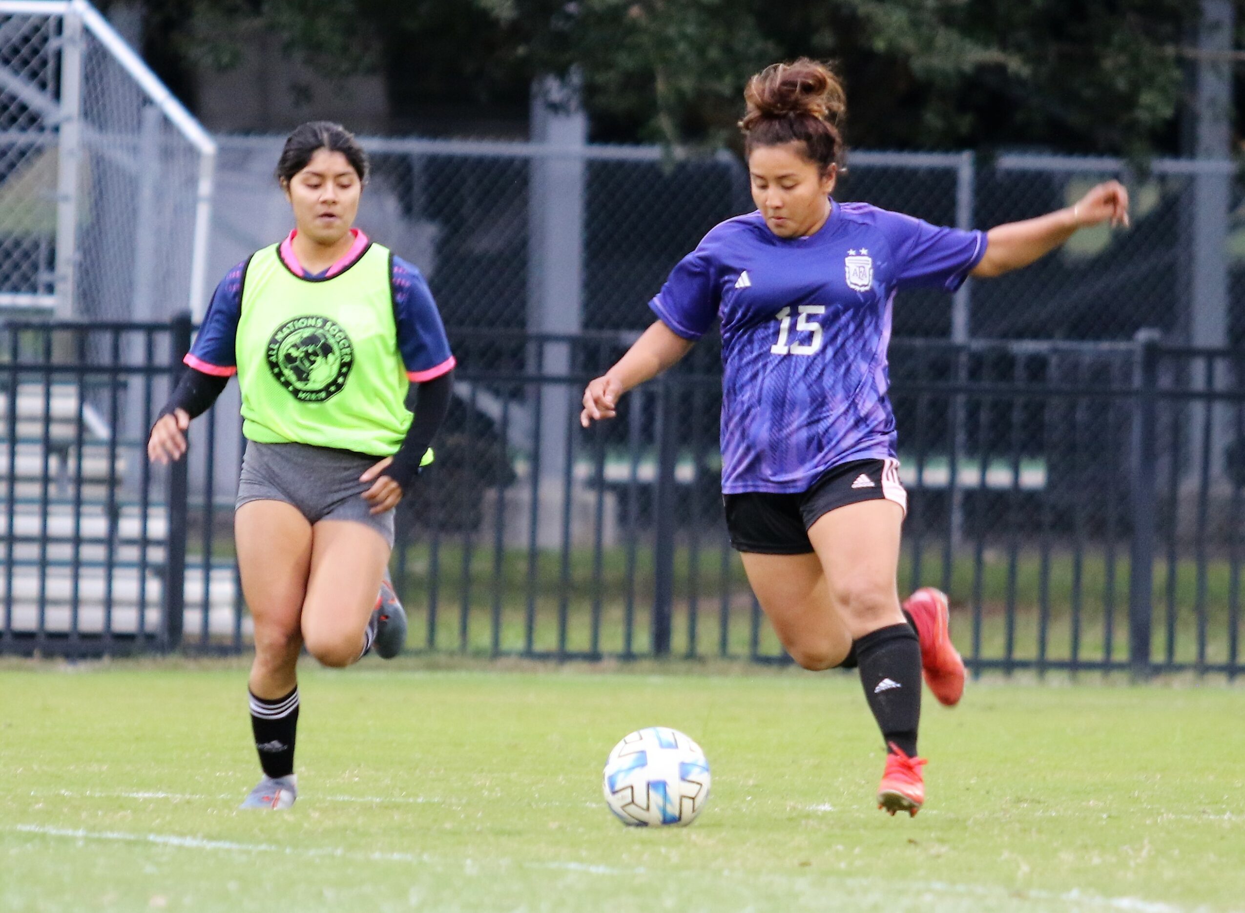 Two female soccer players on a field, one in a blue jersey kicking a ball, the other in a green vest running nearby.