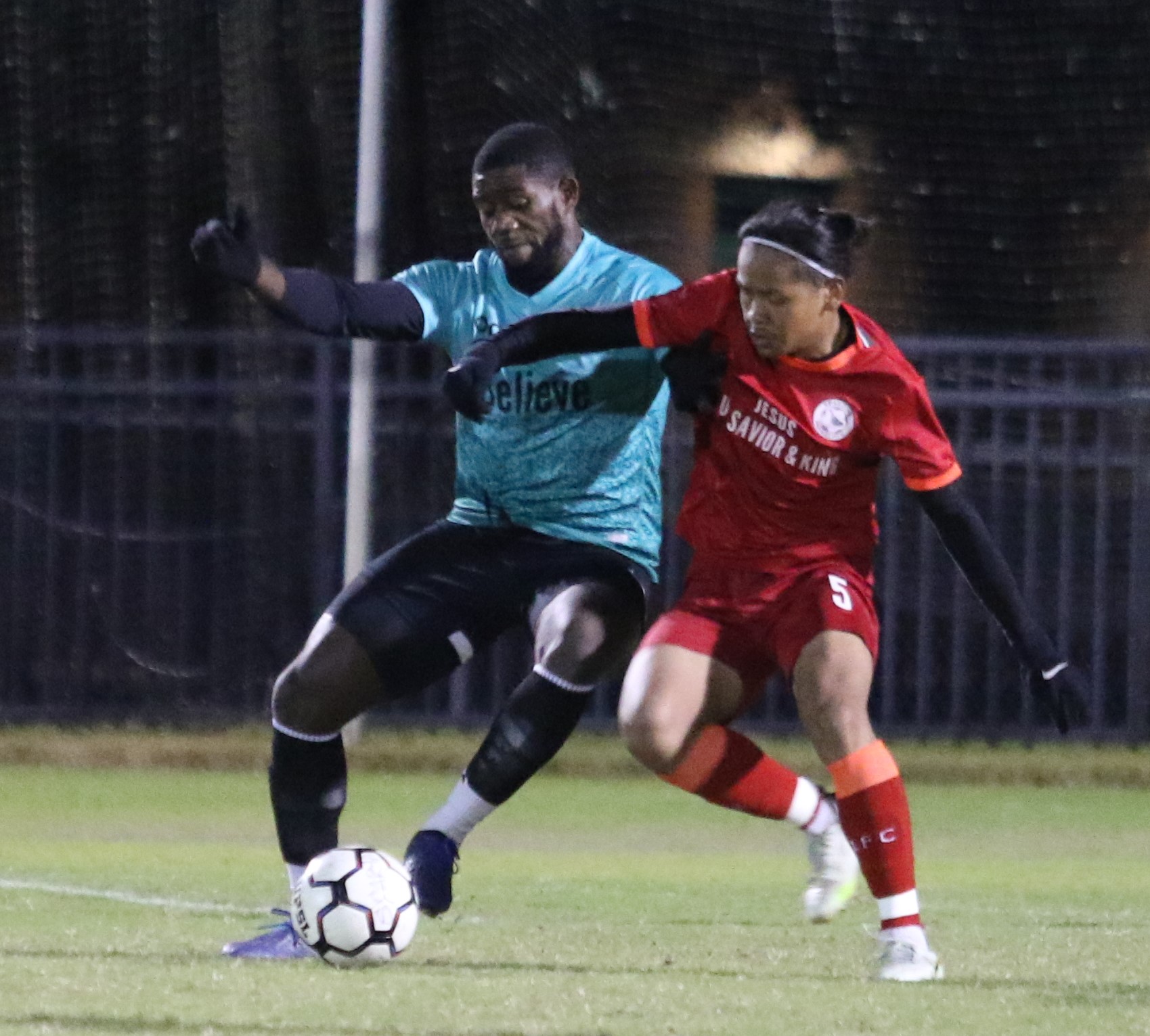 Two soccer players compete for the ball on a field at night, one in a blue jersey and the other in red.