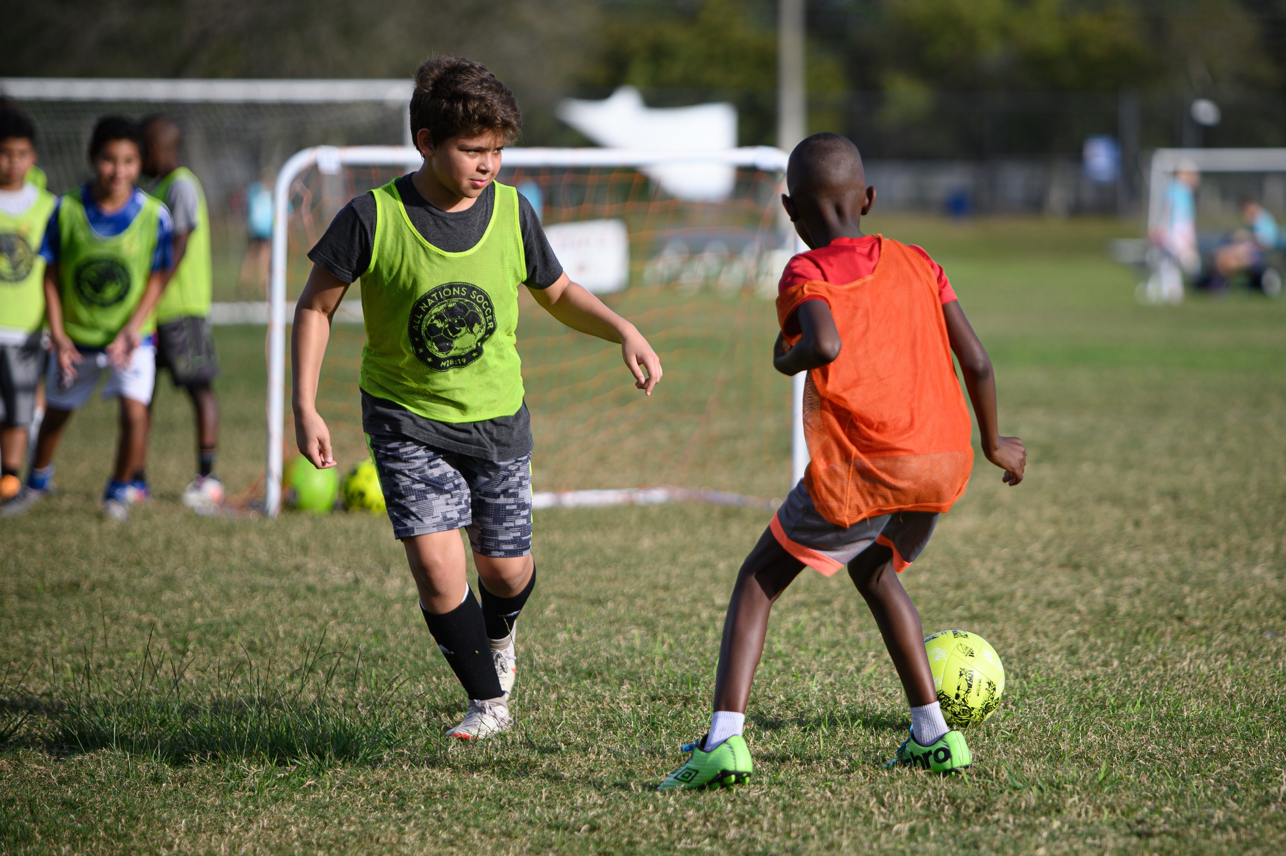 Children playing soccer on a field, with one child in an orange vest and another in a green vest, others in the background.