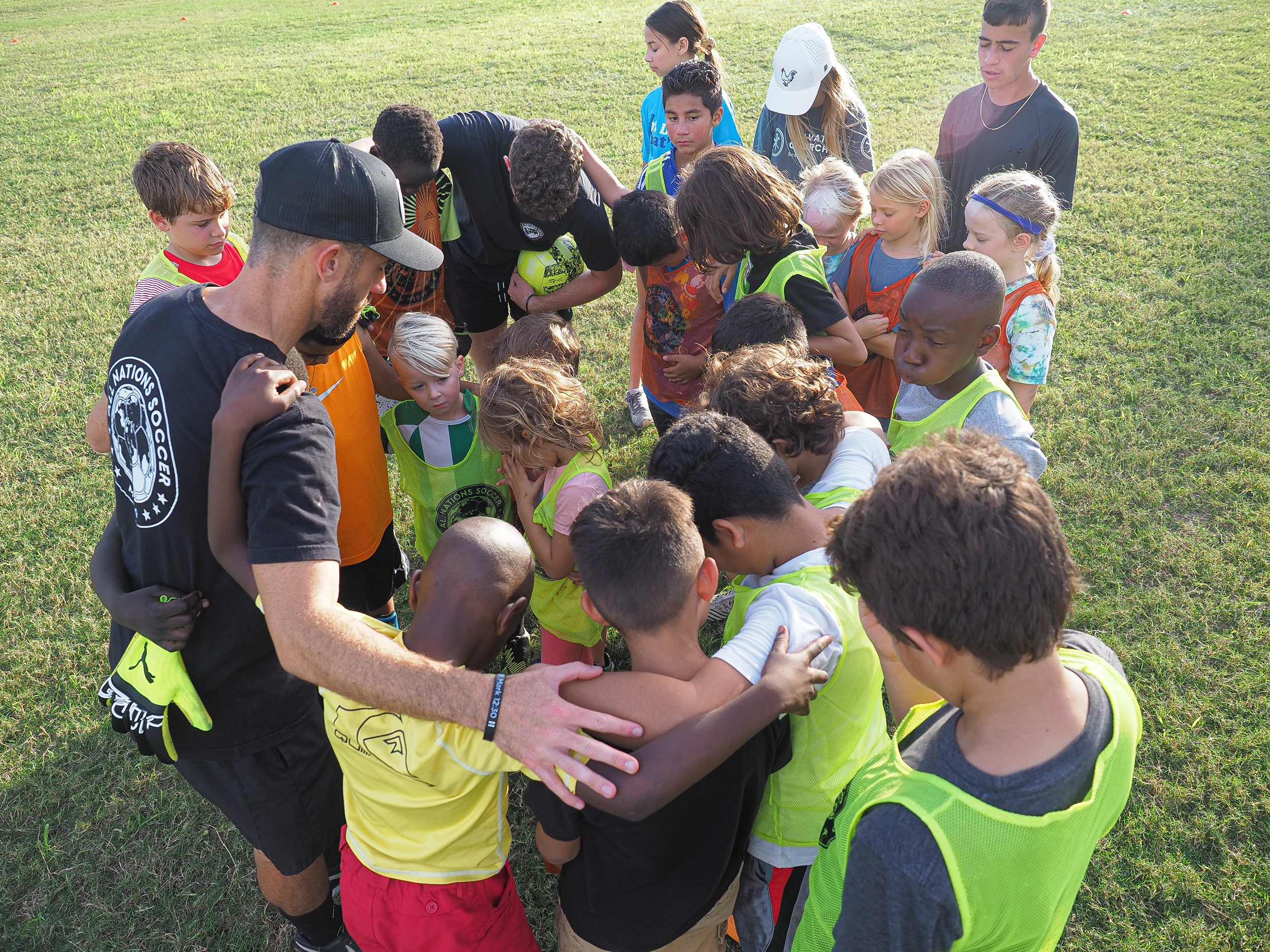 Group of children and adults gathered outdoors on grass, engaging in a group activity or discussion.