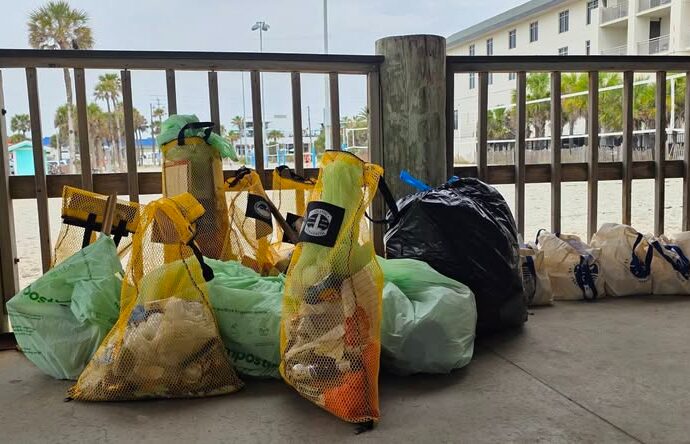 Bags of trash and cleaning supplies lined up against a wooden fence near a beachside area.