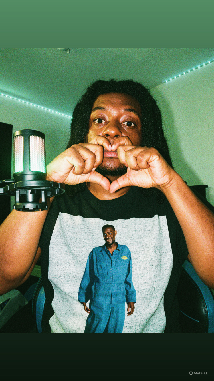 Person with long hair making a heart shape with hands, wearing a T-shirt with a man in a blue suit printed on it, in a room with green walls and LED lights