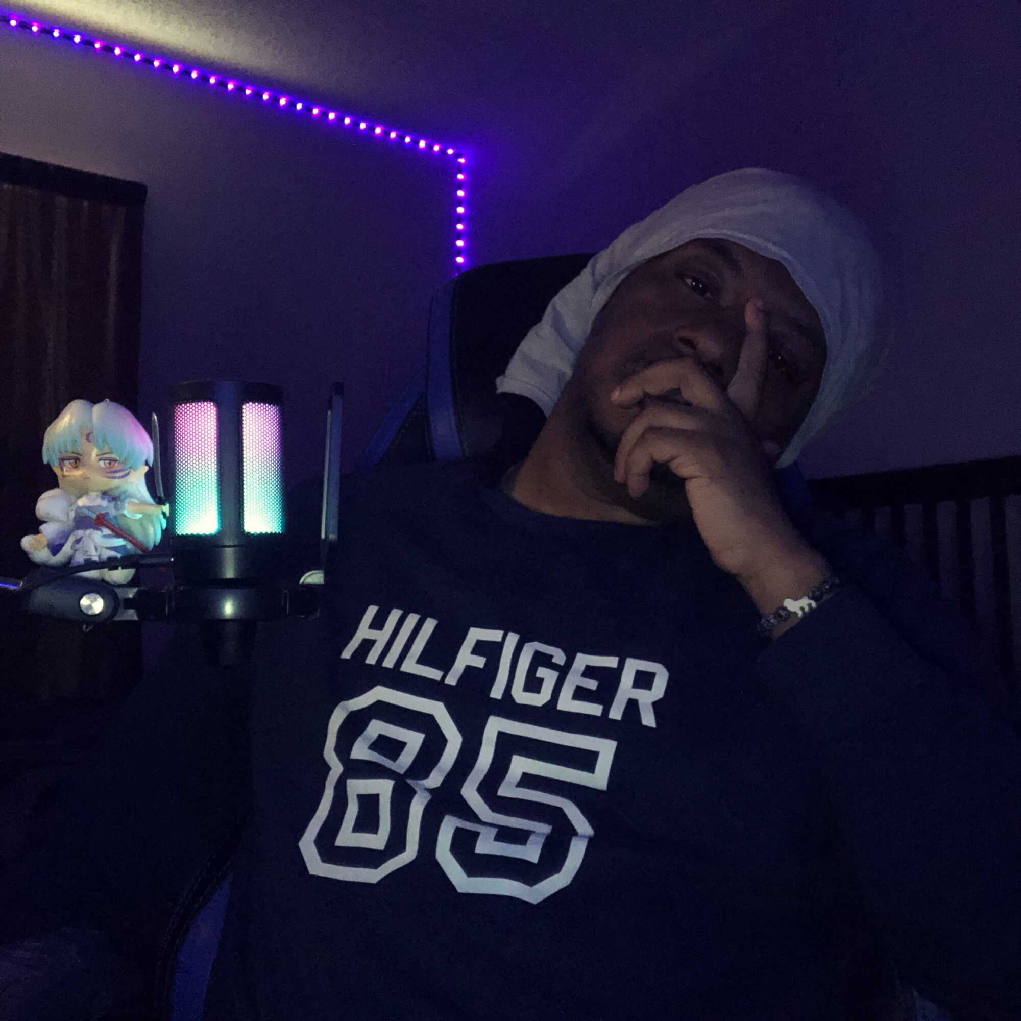 Person wearing a white head covering and a dark shirt with 'HILFIGER 85' text, sitting in a dimly lit room with colorful LED lights.
