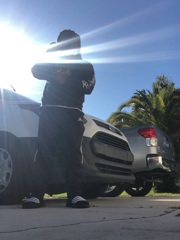 Person standing outdoors near parked cars, with sunlight behind them and trees in the background.