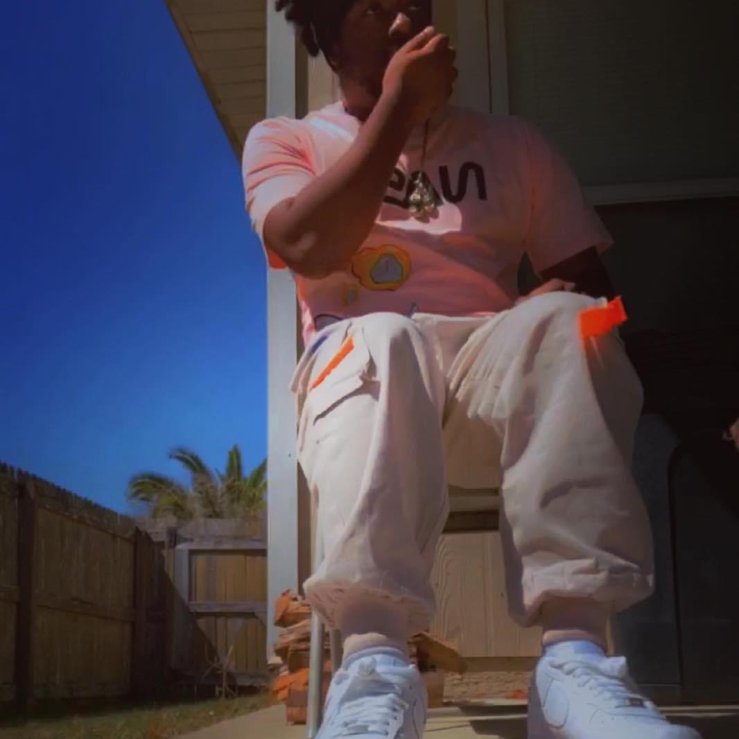 Child sitting outdoors, wearing white pants and sneakers, with a blue sky and palm trees in the background.