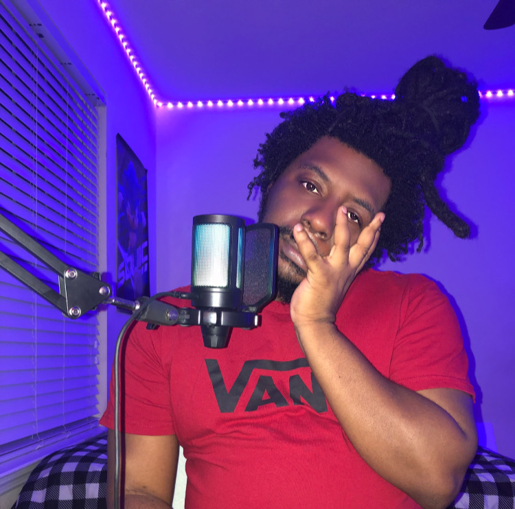 Person with curly hair in a bun, wearing a red Vans shirt, sitting in a room with purple LED lights, near a microphone.