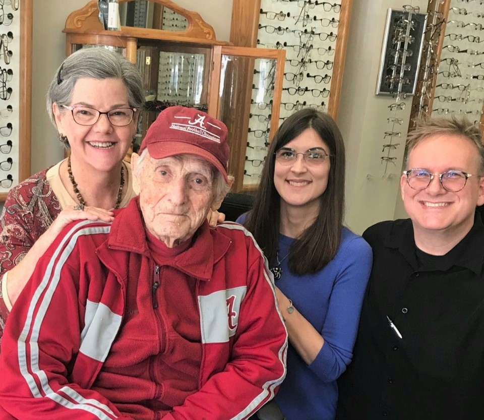 Four people, including an elderly person in a red jacket and cap, smiling indoors with eyeglasses and a mirror behind.