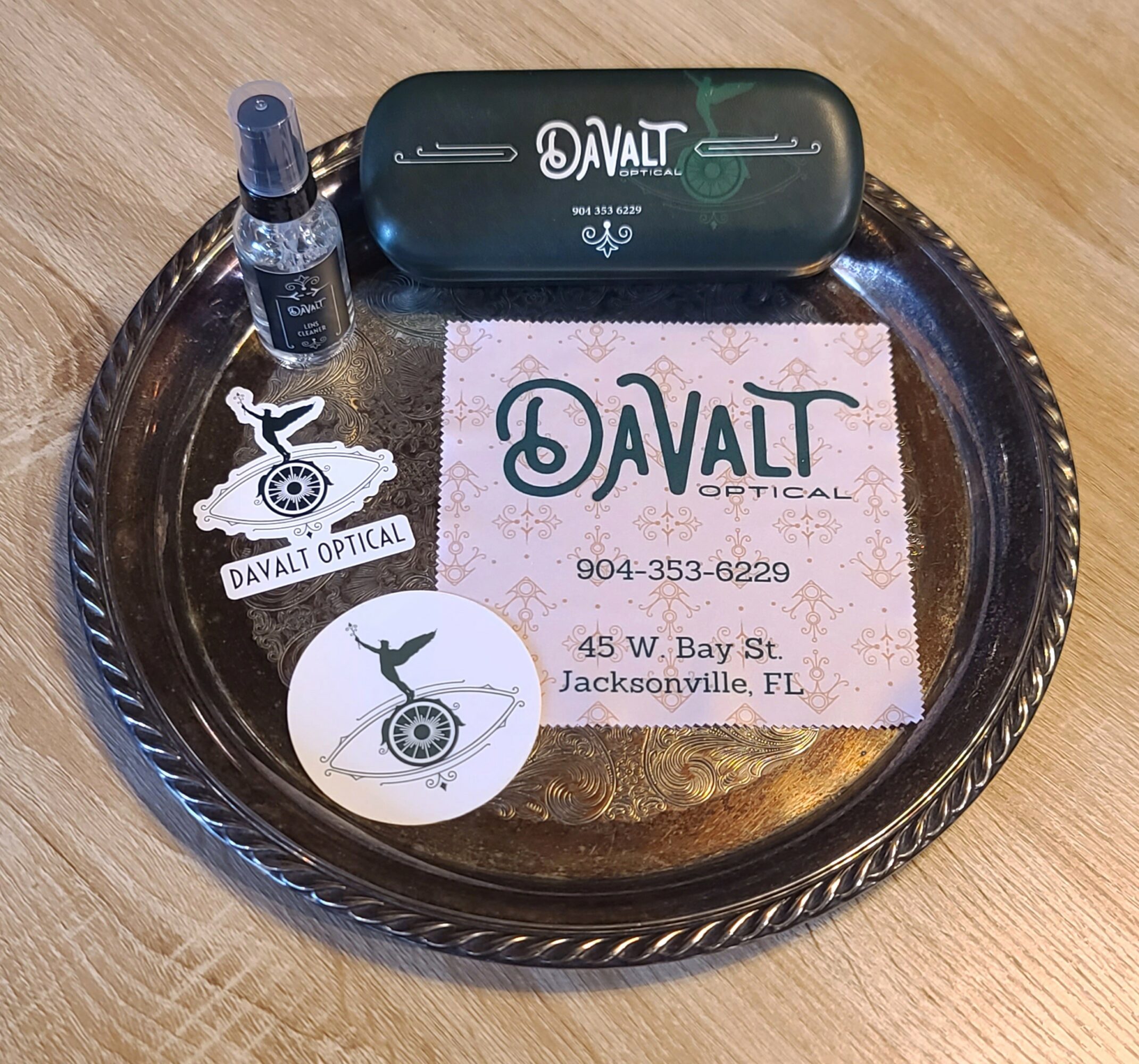 Tray with eye drop bottle, eyeglasses case, business card, and sticker on wooden surface.