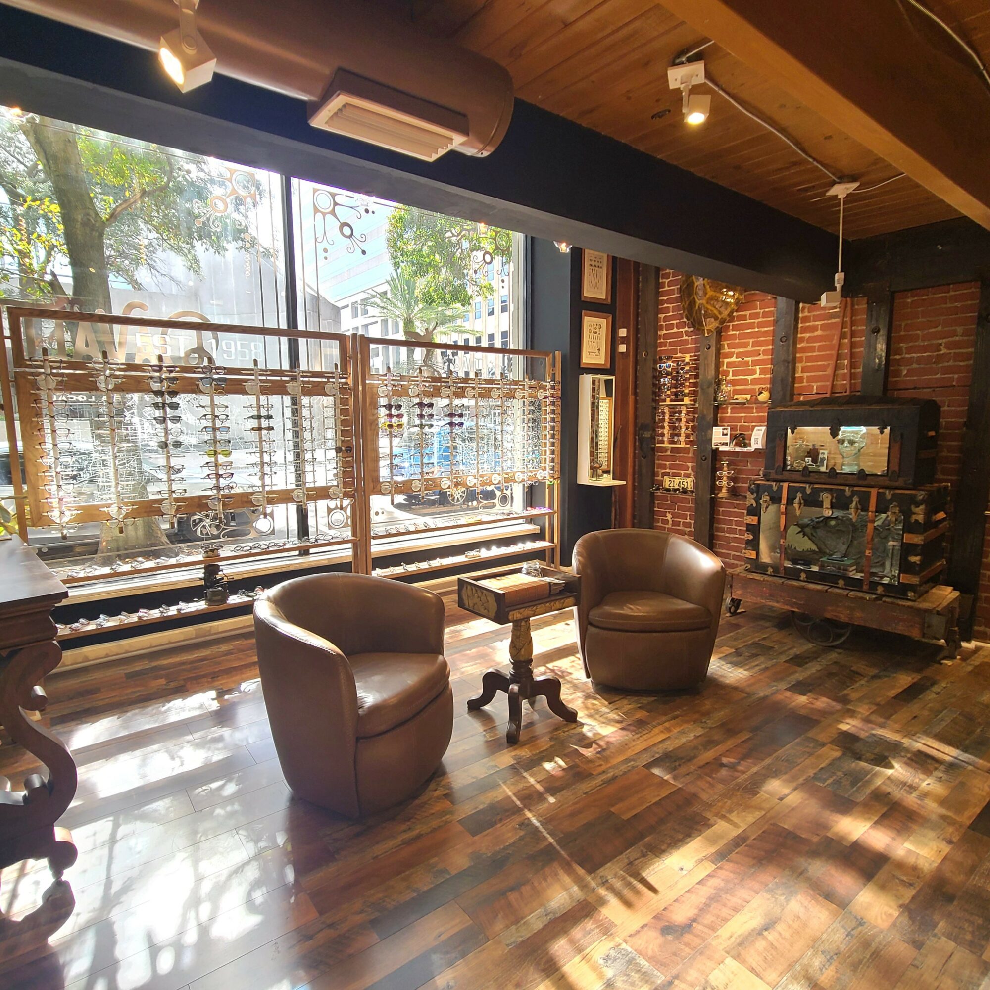 Two leather armchairs with a small table between them in a room with large window, wooden floor, and brick wall.