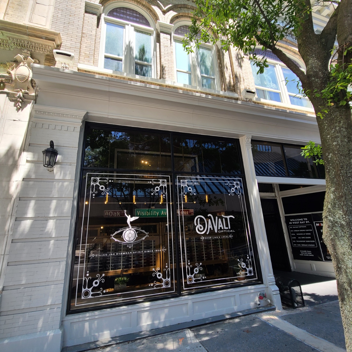 Storefront with large glass windows and decorative white framing, a tree partially obscures the view, and a sign reads 'VALT'.