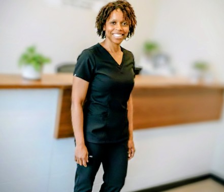 Smiling woman in black scrubs standing in front of a reception desk with plants in the background.