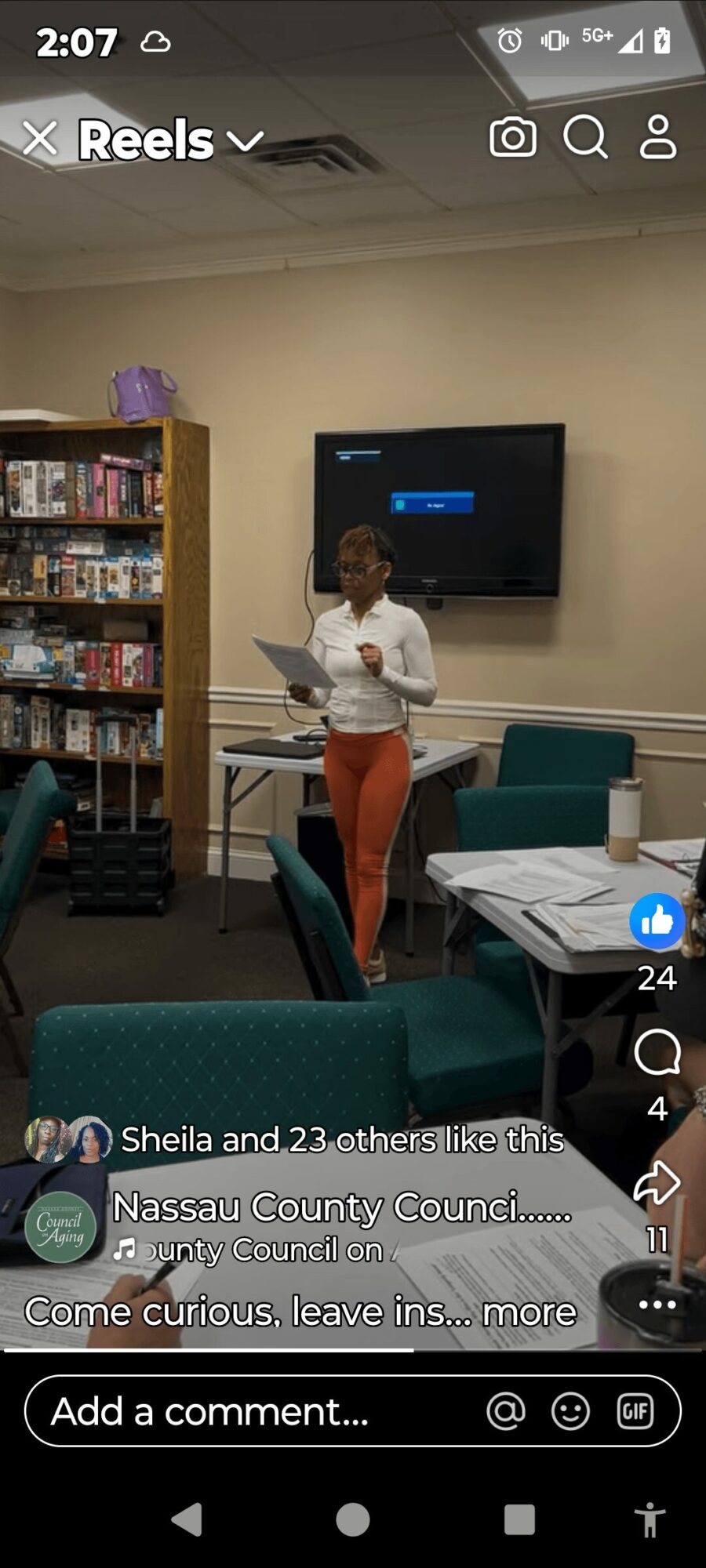 Person standing in front of a TV, holding papers, in a room with bookshelves and tables.
