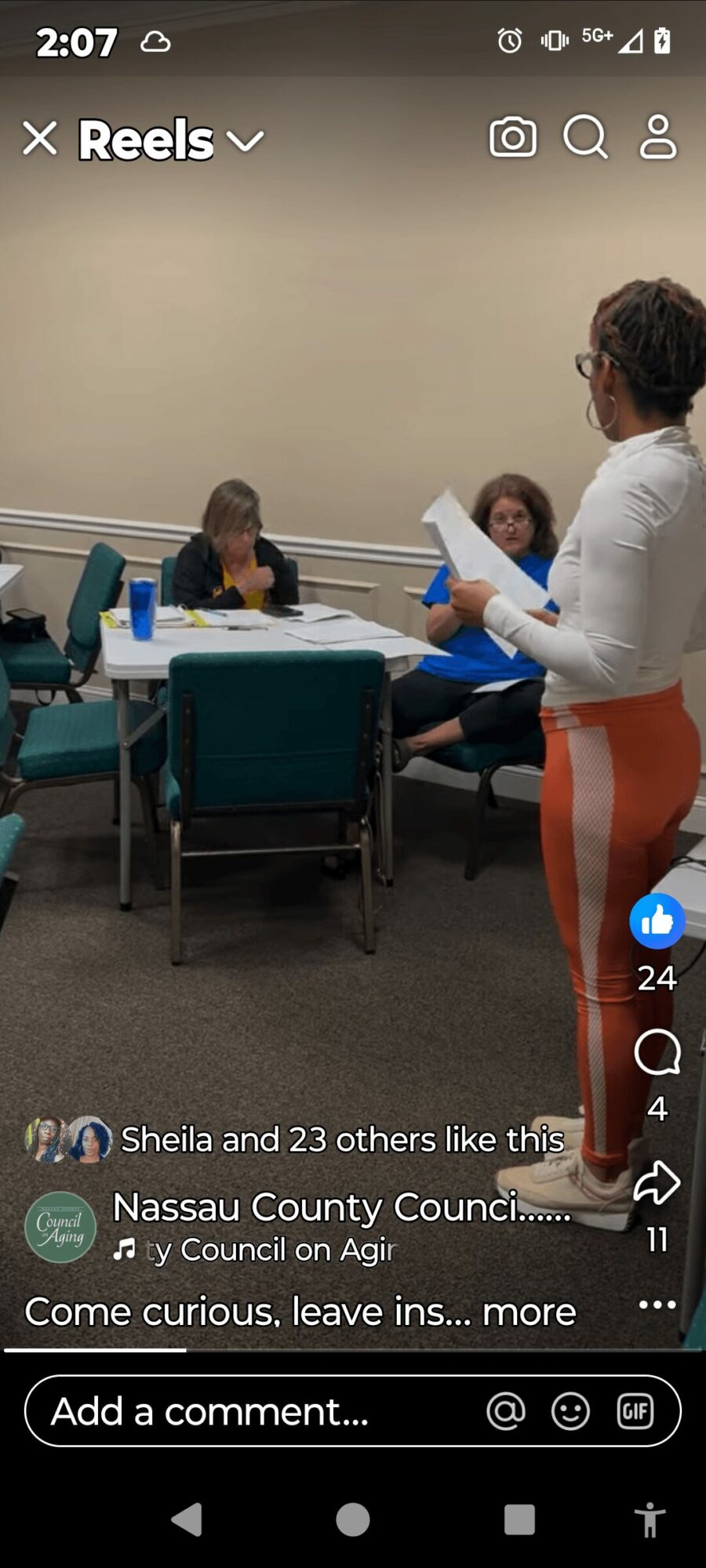 Four people in a meeting room, three seated and one standing, with papers and a water bottle on the table.