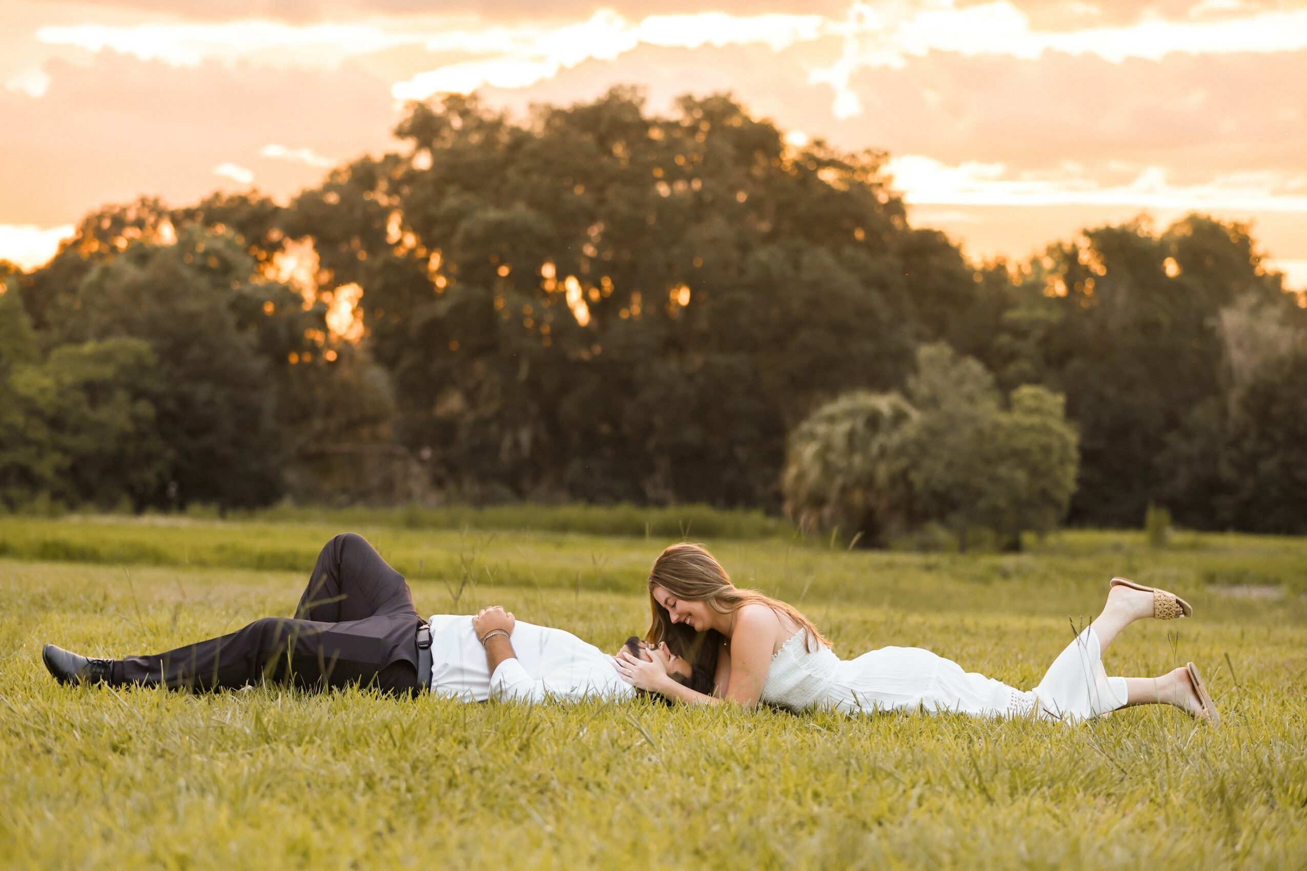 A woman in a white dress lies on her stomach on a grassy field, reading a book, with a man lying beside her.