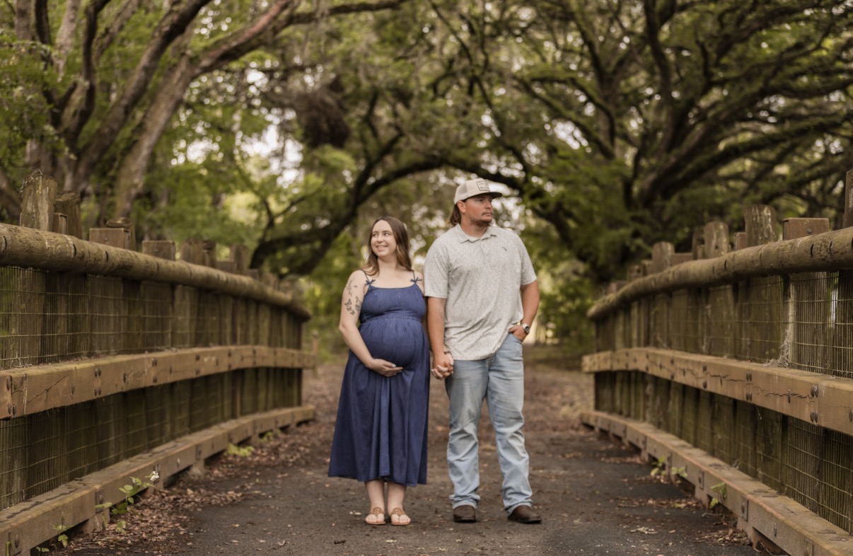A pregnant woman and a man walk together on a wooden bridge surrounded by trees, holding hands and smiling.
