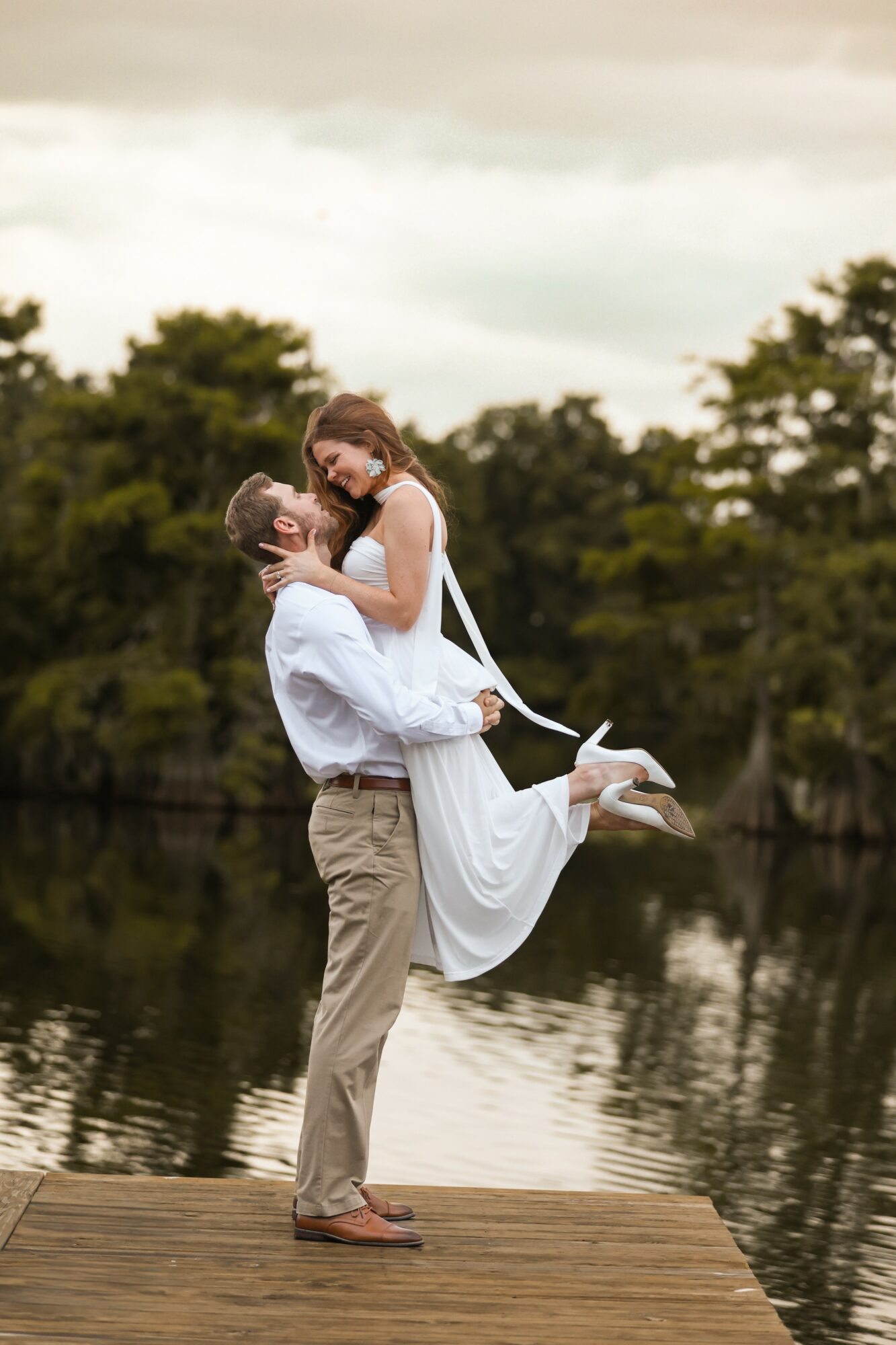 A couple on a wooden dock by a lake, with the man lifting the woman, both smiling, trees in background.