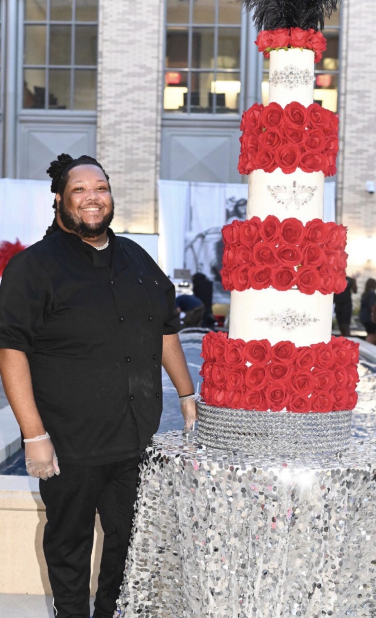Person smiling next to a tall, multi-tiered cake decorated with red roses and silver accents outdoors.