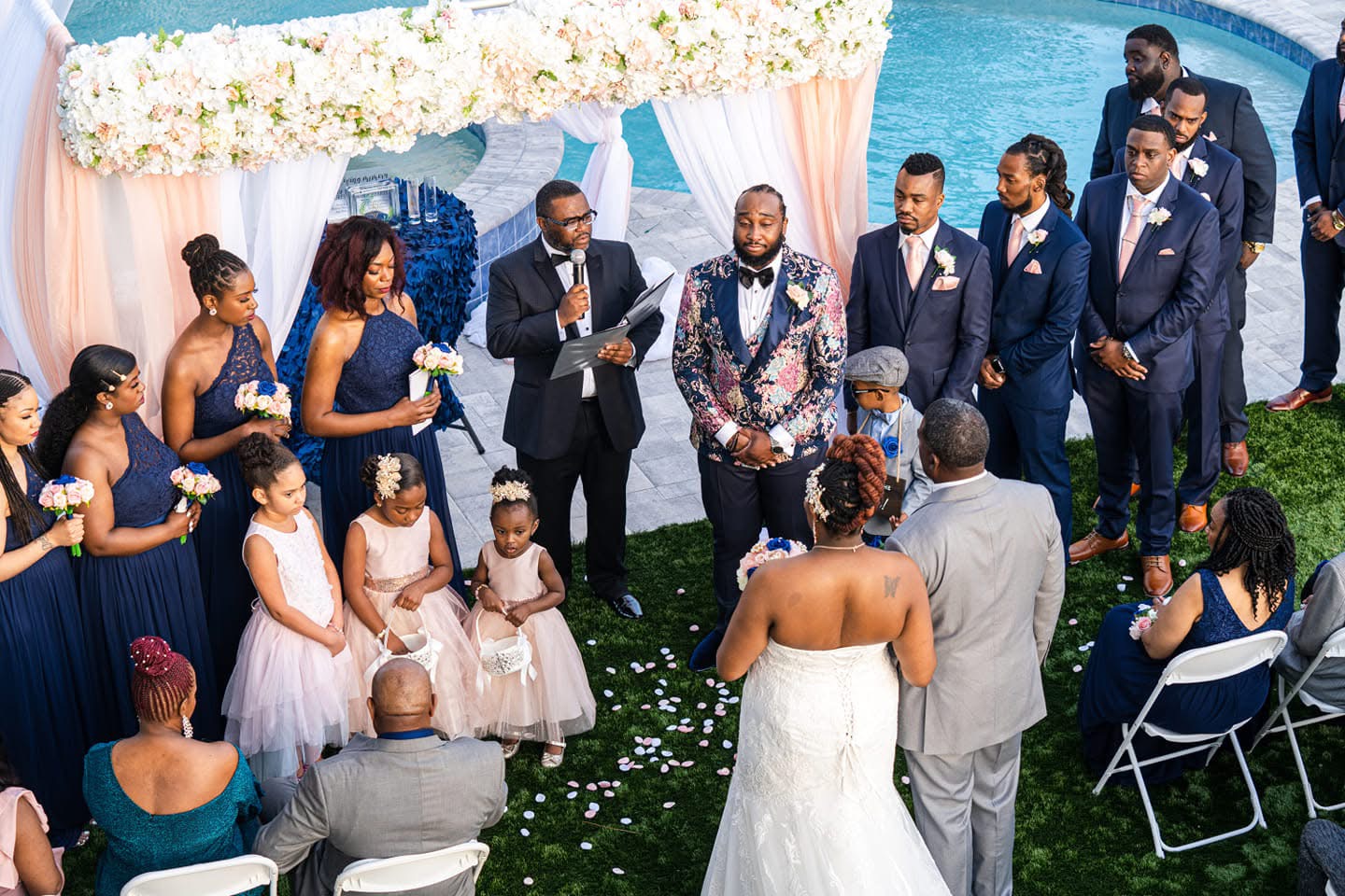 Group of people at a wedding ceremony outdoors, with bridesmaids, flower girls, and groomsmen, under a decorated canopy.