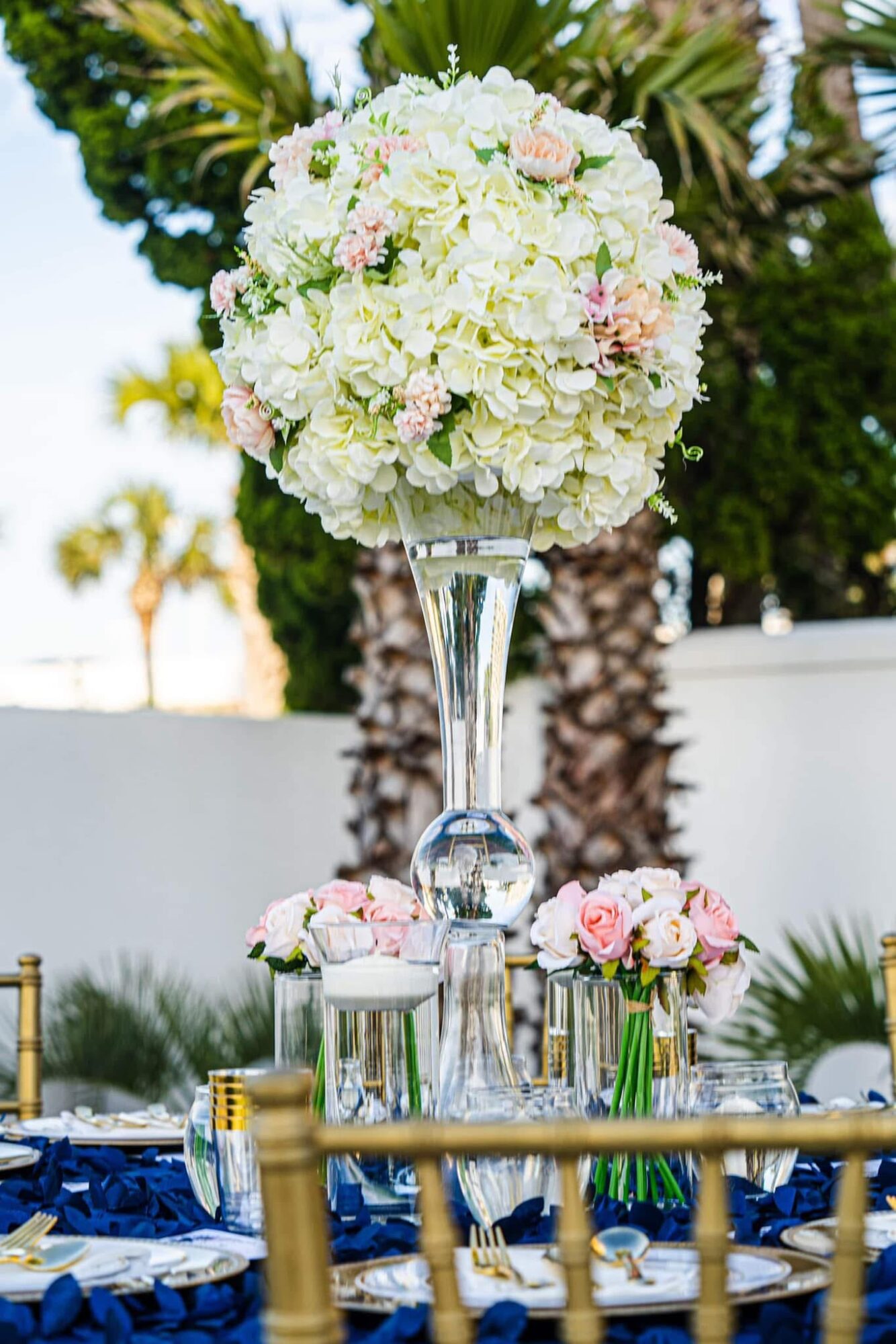 A tall floral centerpiece with white and pink flowers on a table outdoors, with trees and a white wall in the background.