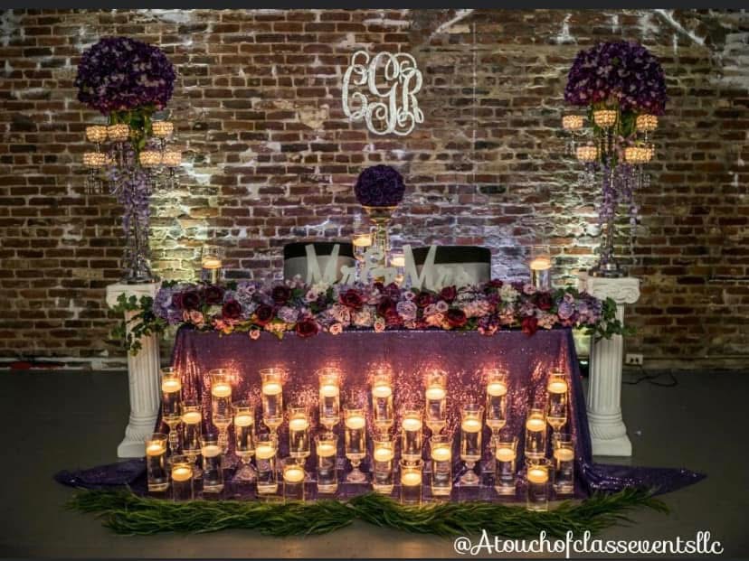 Decorated table with candles, flowers, and a brick wall background, featuring a monogram and floral arrangements.