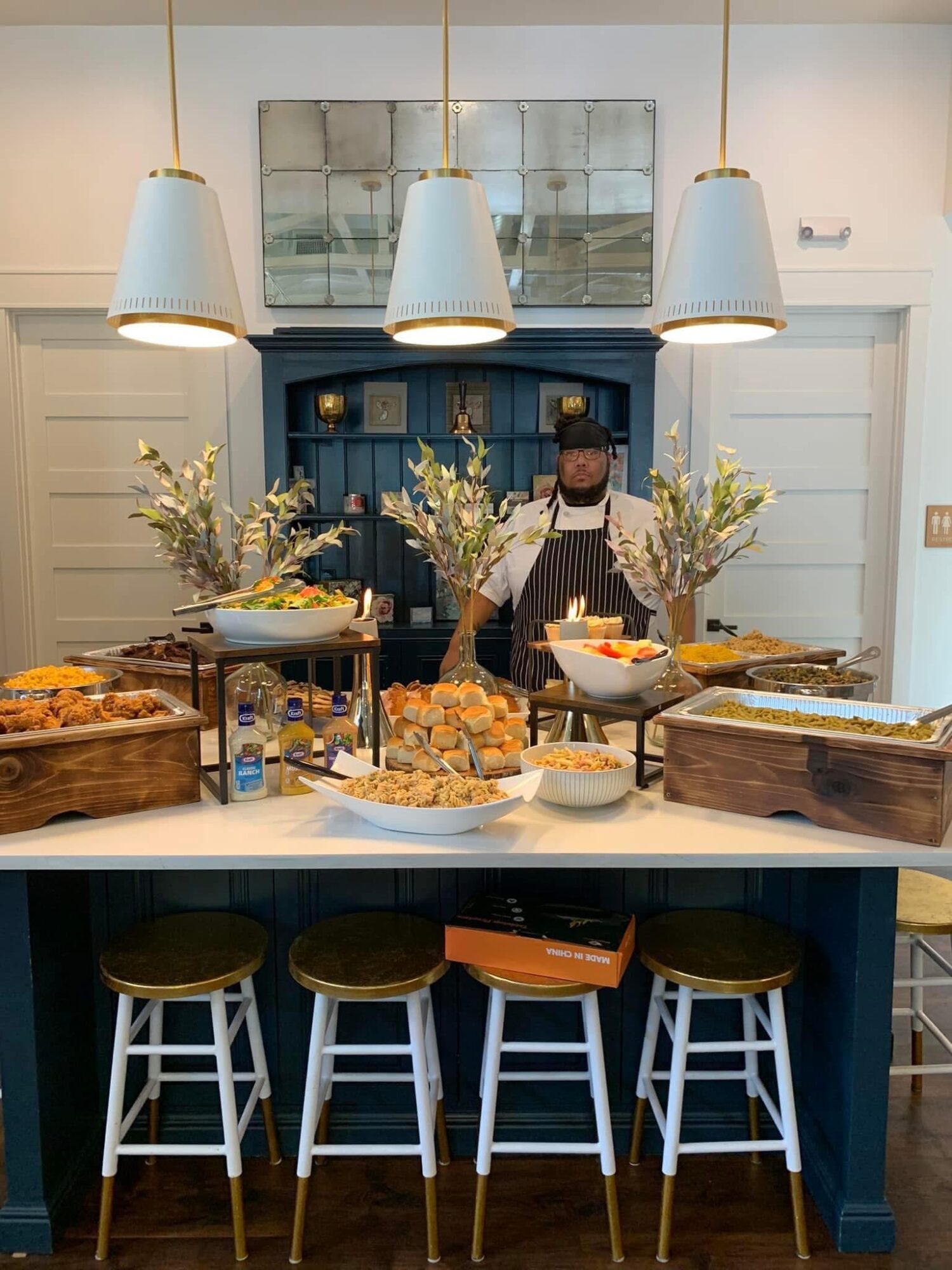 Person behind a buffet table with food, flowers, and three hanging lights in a room with blue walls and white trim.