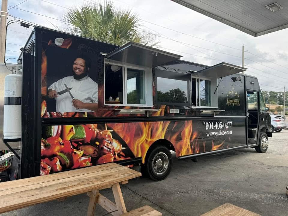 Food truck with a smiling chef, colorful food images, and contact information, parked outdoors with a wooden bench nearby.